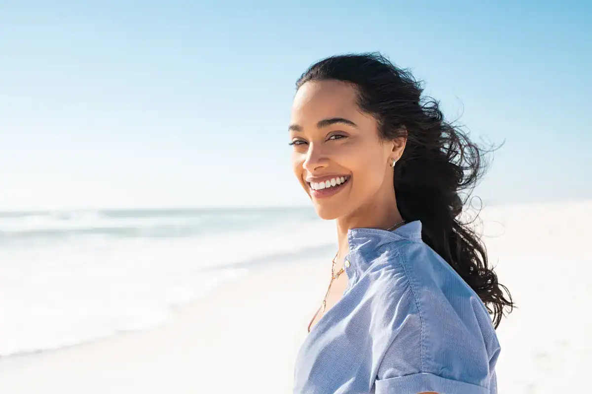 A smiling woman at the beach with clear skin and dark curly hair, sunny day.